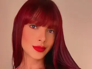 TatyBrazilian poses with long red hair and red lipstick, looking directly at the camera against a plain background.