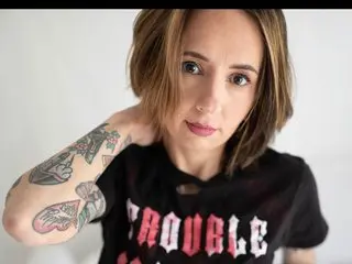 TaylaRae poses with her hand in her hair, wearing a black graphic t-shirt against a plain white background.