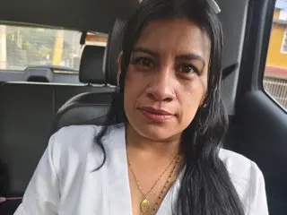 Smiling woman in a white blouse and gold necklace sitting in a car seat on a bright day.