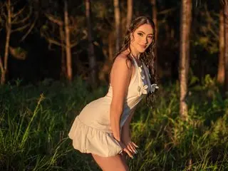 TaylorAdaams poses in a white sundress against a soft, sunlit forest background, showing her long brown hair and bare shoulders.