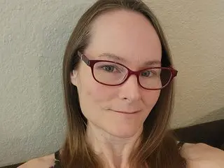 TeamJerica poses with a slight smile, wearing red-rimmed glasses and showing her long brown hair against a plain wall.