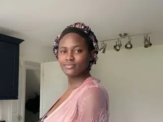 Tempting_Aura poses with a soft smile, wearing a pink lace top and a dark patterned headwrap against a plain wall.