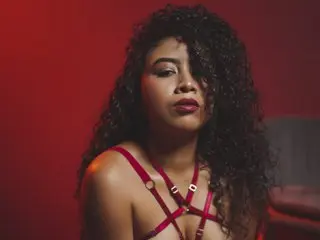 Tharaa24 poses with long curly hair wearing a red strappy harness against a red background.