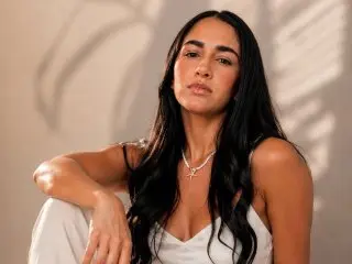 ThayssaRoss poses with long dark hair, wearing a white tank top against a neutral background.