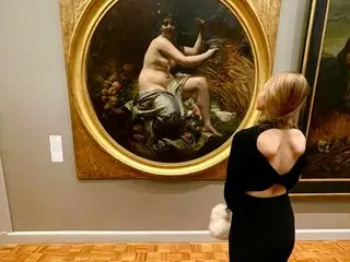 Blonde woman in a black backless dress admiring a classical gold-framed painting in an art museum gallery.