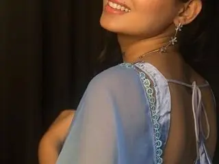 TheSassygirl smiles while wearing a light blue sheer top that shows her shoulders and neck against a dark background.