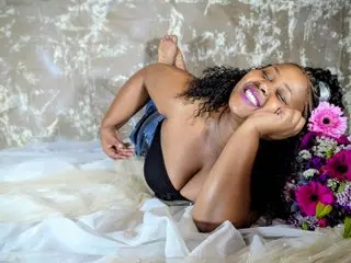 Smiling Black woman in black lingerie lying on white fabric next to pink flowers in a dreamy setting.