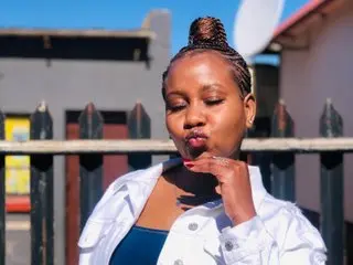 Thickleex poses outdoors with her hair in a high bun, wearing a white button-down shirt and a blue top.
