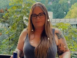 Thickthighsbig poses with long brown hair, wearing a grey tank top, while standing outdoors in front of green trees.