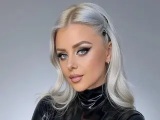 TiffanyDeen poses with her long silver hair and heavy eye makeup, wearing a black shiny latex top against a plain grey background.