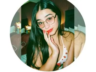TinaClaytoon wearing glasses and a white floral bikini top, posing with her hand on her chin in a bedroom.