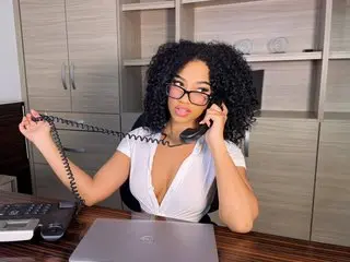 TinaHarris with curly black hair wearing a white tank top, holding a black telephone receiver to her ear while sitting at a desk.
