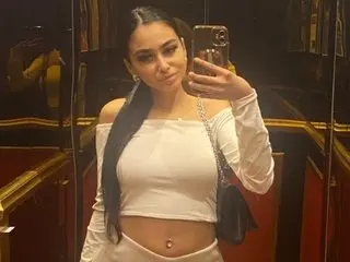 TinyCassy poses in a white long-sleeve crop top, showing her long dark hair and midriff in a dimly lit room.