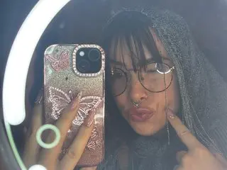 TinytittieXX poses for a mirror selfie, wearing a dark top and showing her dark hair and face with glasses.