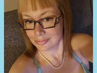 Smiling woman with blonde bangs and glasses wearing a pearl necklace and light blue strap top indoors.