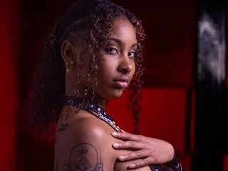 Triipsy poses with her hands on her chest, showing her curly hair, facial features, and a black strap or top against a dark red background.