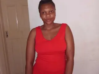 TyraTrixie poses in a red tank top against a plain white wall, showing her bare arms and shoulders.