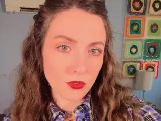 Unicornfairy55 looks directly at the camera with long wavy brown hair and red lipstick, wearing a blue and white plaid shirt.