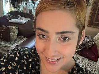UnitedTrees smiles close up, showing her short blonde hair and wearing a black floral print top while sitting on a brown couch.