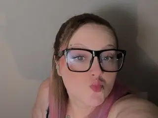 VELVETDesires poses close-up, wearing black glasses and showing her face and shoulders against a plain grey background.