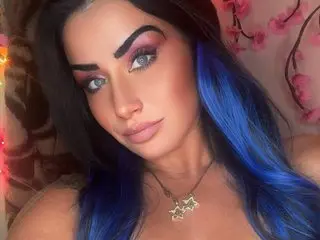 ValayaNoire poses with long blue hair and heavy eye makeup, wearing a gold necklace against a soft pink floral background.