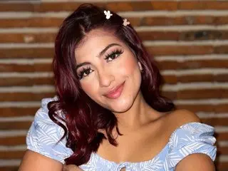 ValeSandoval poses with her long dark hair and a light blue off-the-shoulder top against a warm, brick-patterned background.