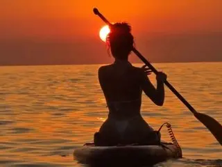 Silhouette of a woman paddleboarding on a calm ocean during a vibrant orange sunset.