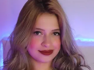 Valeria_Estevez poses with a smile, showing her long blonde hair and light makeup against a soft purple background.