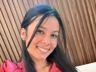 ValerieNoiir smiles while looking at the camera, showing her long dark hair and wearing a red top against a wooden slatted wall.