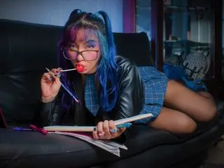 ValeryTatto poses in a black leather jacket and blue plaid skirt, showing off her blue-tinted hair and glasses while sitting on a black leather sofa.