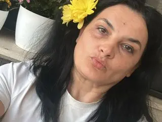 Smiling woman with black hair and a yellow flower in her hair wearing a white t-shirt in a sunny garden setting.