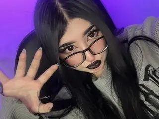 Goth girl with long black hair and glasses wearing a grey sweater in moody purple lighting.