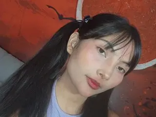 Vanelovx poses with long black hair and bangs, looking directly at the camera against a warm orange background.