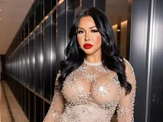 VanessaBacherin poses with long dark hair, wearing a sheer, sparkling tan lace bodysuit that shows her cleavage and skin.