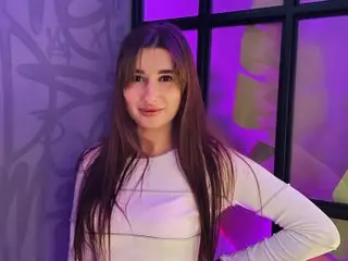 Vanila_A poses with long brown hair, wearing a white long-sleeved shirt against a purple and black background.