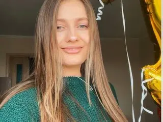 VasilisaRox smiles with her long blonde hair down, wearing a green textured top against a plain indoor background.