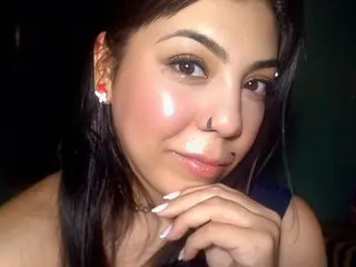 Vecina_Caliente looks at the camera with long dark hair and a slight smile, showing her face and shoulders against a dark background.