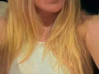 VeeBeeXoX with long blonde hair, wearing a white tank top, posing against a soft-focus background.