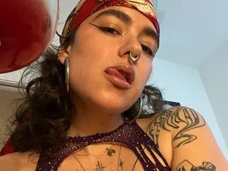 Velvette_Peach poses close-up, showing her face, dark wavy hair, and nose ring while wearing a red patterned headwrap and purple beaded jewelry.