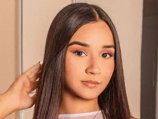 Young woman with long straight dark hair and soft glam makeup posing in a bright indoor setting.