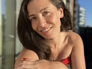 VerityDawnUK smiles while posing with her long brown hair down, showing her bare shoulders and collarbone in a bright, sunlit setting.