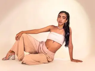 Veronica_Hottiee poses in a white tube top and tan trousers while leaning back on a white floor.