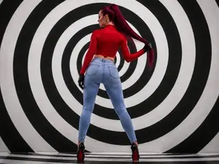 VictoriaHeller stands with her back to the camera, wearing a red long-sleeve top and light blue jeans against a black and white spiral background.