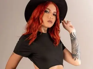 VictoriaRossX poses with red hair and a black hat, wearing a black crop top that shows her neck and shoulders against a plain background.