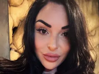 VictoriaSummersUK poses close-up, showing her face, dark wavy hair, and pink lips against a warm, textured background.