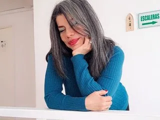 VikyAdams72 with long grey hair leans her chin on her hand while wearing a blue long-sleeved ribbed top.