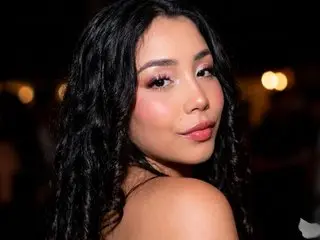 Young woman with wavy black hair and glowing makeup smiling in a dimly lit evening setting.