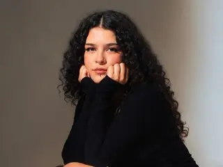 VioletSing poses with her hands under her chin, wearing a black long-sleeved top and letting her long, dark curly hair hang down.