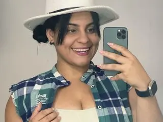 Violethrousse smiles while posing in a white hat and a blue and white plaid shirt, holding a smartphone to take a mirror selfie.
