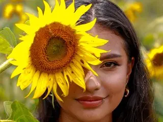 ViolettaK poses with a large sunflower partially covering her face, showing her dark hair and smile in an outdoor setting.